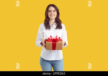 Happy brunette woman in white shirt is holding a present gift in hands on yellow background. Stockfoto