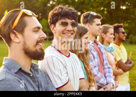Fröhlicher junger Mann, der mit seinen Freunden in Reihe steht und während des Sommerparks Händchen hält. Stockfoto