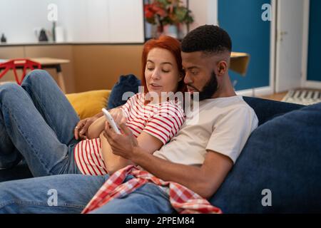 Junge, vielseitige Paare, die sich zu Hause mit dem Mobiltelefon ausruhen Stockfoto