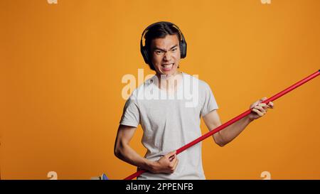 Asiatischer Sorgenloser, der Gitarre mit Besen spielt, sich vor der Kamera lustig macht und Musik hört. Fröhlicher, lustiger Mann, der lächelt und Lieder singt, ein Audio-Headset benutzt, Spaß mit Geräuschen zur Unterhaltung hat. Stockfoto