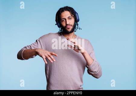 Selbstbewusster arabischer Mann macht coole Bewegungen, während er Musik über Kopfhörer hört und in die Kamera schaut. Junge Person tanzt, während sie Musik im Kopfhörerporträt hört Stockfoto