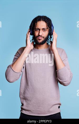 Ein cooler arabischer Mann, der Musik hört, während er Kopfhörer am Kopf berührt und in die Kamera schaut. Selbstbewusste Person mit ernsthaftem Gesichtsausdruck, die Musik im Kopfhörerporträt genießt Stockfoto