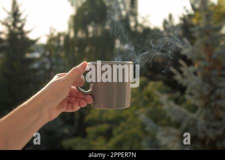 Die Hand einer Frau hält eine Tasse Kaffee vor dem Hintergrund der aufgehenden Sonne. Nahaufnahme. Heißes Getränk in einer Tasse und auf einer Tasse Stockfoto