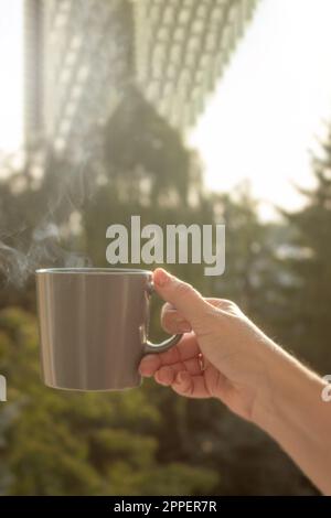 Die Hand einer Frau hält eine Tasse Kaffee vor dem Hintergrund der aufgehenden Sonne. Nahaufnahme. Heißes Getränk in einer Tasse und auf einer Tasse Stockfoto