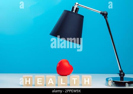 Rotes Herz auf Holzwürfeln mit dem Wort GESUNDHEIT auf einem Tisch neben einer schwarzen Tischlampe Stockfoto