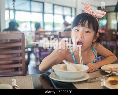 Glückliches asiatisches Kind, das im Restaurant leckere Nudeln mit Stäbchen isst Stockfoto