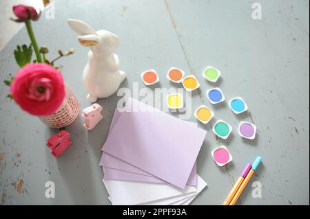 Viele Handwerksvorräte auf einem Holztisch. Papier, Marker und farbige Blätter auf einem Tisch, dekoriert mit Blumen und Kaninchen. Stockfoto