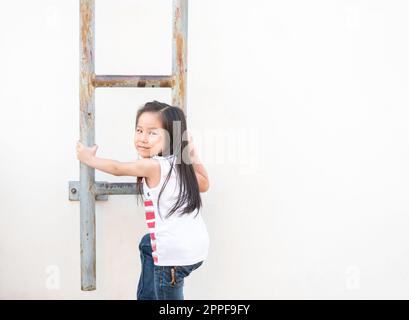 Ein kleines asiatisches Mädchen klettert die Treppe hoch - Feuerleiter, der weiße Wandhintergrund Stockfoto