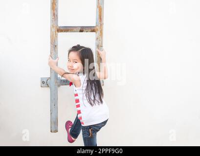 Ein kleines asiatisches Mädchen klettert die Treppe hoch - Feuerleiter, der weiße Wandhintergrund Stockfoto