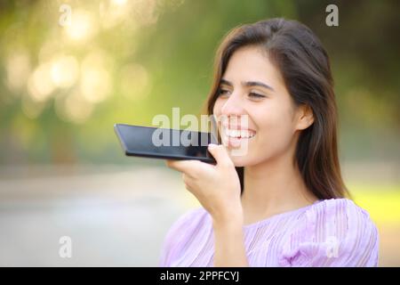 Glückliche Frau, die in einem Park Spracherkennung am Handy benutzt Stockfoto