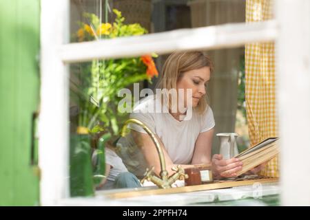 Eine Mid-Frau sieht sich ein Rezeptbuch an einem offenen Fenster in der Küche an Stockfoto