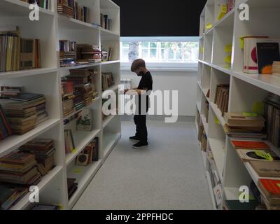 Sremska Mitrovica, Serbien 15. Mai 2021 ein 8-jähriger Junge mit Maske und schwarzem T-Shirt steht neben dem Bücherregal in der Bibliothek und liest ein Buch. Schulbildung. Liebe für Literatur und Lesen Stockfoto