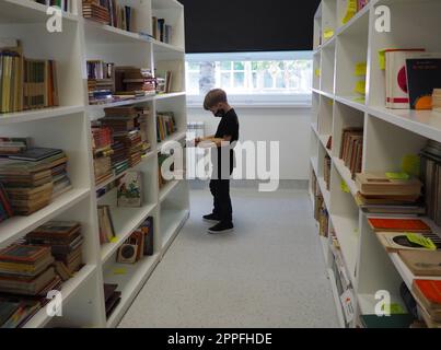 Sremska Mitrovica, Serbien 15. Mai 2021 ein 8-jähriger Junge mit Maske und schwarzem T-Shirt steht neben dem Bücherregal in der Bibliothek und liest ein Buch. Schulbildung. Liebe für Literatur und Lesen Stockfoto