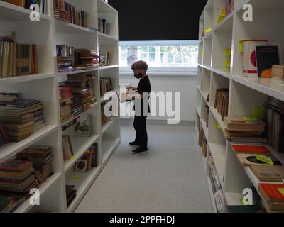 Sremska Mitrovica, Serbien 15. Mai 2021 ein 8-jähriger Junge mit Maske und schwarzem T-Shirt steht neben dem Bücherregal in der Bibliothek und liest ein Buch. Schulbildung. Liebe für Literatur und Lesen Stockfoto