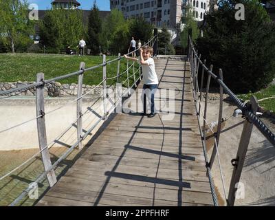 Hängende Holzbrücke. Der Junge versucht, die Brücke zu rocken. Stanisici, Bijelina, Bosnien und Herzegowina. Eine Brücke aus Brettern und Geländern, die von 4 Personen getragen werden kann. Stockfoto