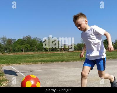 Ein Junge läuft einem Basketballball hinterher. Das Kind spielt mit einem Ball auf dem Spielplatz. Geben Sie Platz für Text frei. Blauer Himmel im Hintergrund. Ein blondes Kind Stockfoto