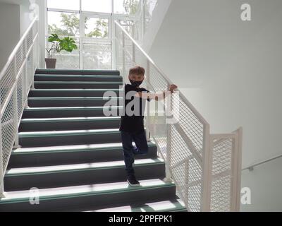 Ein blonder Junge, 8 Jahre alt, in schwarzen Kleidern und einer Maske, steht auf einer weißen Schulleiter. Das Kind schaut in die Kamera und hält den Handlauf mit seiner Hand. Schulkonzept. Innenraum. Stockfoto