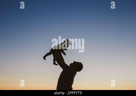 Silhouette von Dad, der das Baby bei Sonnenuntergang in der Luft hält Stockfoto