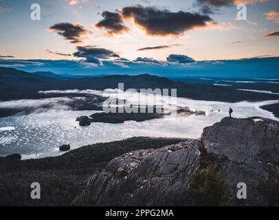 Ein einsamer Wanderer steht auf dem felsigen Gipfel mit Seen und Bergen, Sonnenaufgang Stockfoto