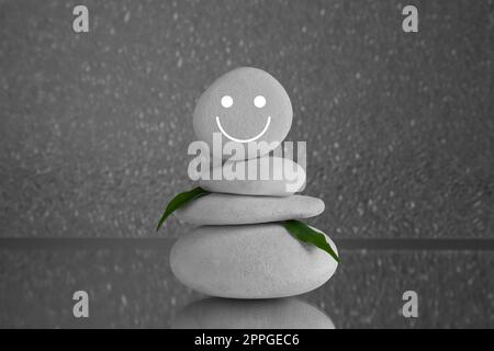 Steine mit gezogenem fröhlichem Gesicht und grünen Blättern auf grauem Hintergrund. Zen-Konzept Stockfoto