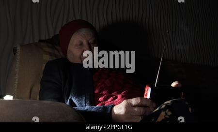 Ukraine: Großmutter hält während russischer Terroranschläge Radio in den Händen, während sie im Dunkeln des Blackouts sitzt. Seniorin in Winterkleidung in Dunkelheit auf dem Sofa mit Handfunkempfänger, der Nachrichten hört Stockfoto