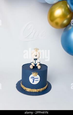 Ein blauer Kuchen mit einem Teddybär oben drauf, mit einer Krone drauf. Kyrillische Inschrift "Misha ist 1 Jahre alt". Kuchen auf weißem Hintergrund Stockfoto