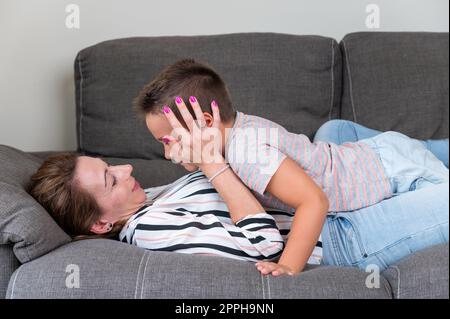 Eine Frau, die auf dem Sofa liegt und ihren kleinen Sohn in die Wange küsst. Mutter und kleiner Junge verbringen fröhlich Zeit im gemütlichen Wohnzimmer zu Hause Stockfoto