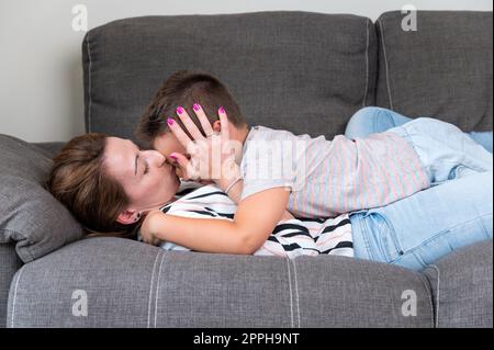 Eine Frau, die auf dem Sofa liegt und ihren kleinen Sohn in die Wange küsst. Mutter und kleiner Junge verbringen fröhlich Zeit im gemütlichen Wohnzimmer zu Hause Stockfoto
