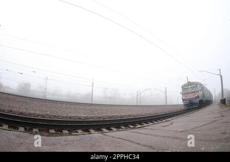 Der ukrainische Vorstadtzug rast an einem nebligen Morgen entlang der Eisenbahn. Fischaugenfoto mit erhöhter Verzerrung Stockfoto