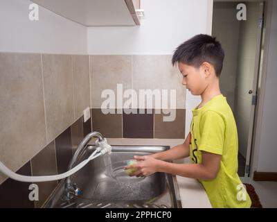 Ein süßer Junge, der Geschirr in der Küche spült. Das Kind hat Spaß dabei, seinen Eltern bei der Hausarbeit zu helfen. Stockfoto