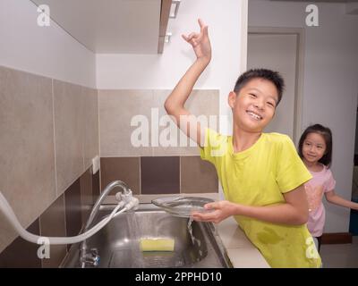 Ein süßer Junge, der Geschirr in der Küche spült. Das Kind hat Spaß dabei, seinen Eltern bei der Hausarbeit zu helfen. Seine jüngere Schwester ist dahinter Stockfoto