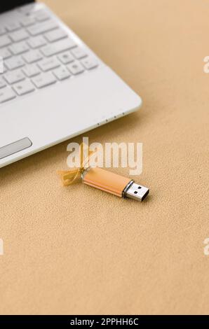 Die orangefarbene usb-Flash-Speicherkarte mit Schleife liegt neben einem weißen Laptop auf einer Decke aus weichem, hellorangefarbenem Fleece. Klassisches weibliches Geschenkdesign für eine Speicherkarte Stockfoto