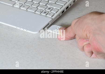 Eine männliche Hand steckt eine weiße kompakte SD-Karte in den entsprechenden Eingang an der Seite des weißen Netbooks. Der Mensch nutzt moderne Technologien, um Speicher und digitale Daten zu speichern Stockfoto