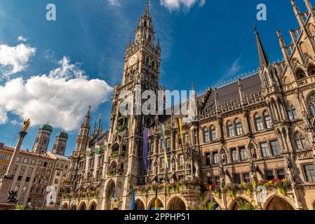 Die neogotische Fassade des neuen Rathauses mit Rathausturm in der Münchner Altstadt im Blick von unten vom Marienplatz bei schönem Sommerwetter und blauem Himmel mit den beiden Türmen der Frauenkirche im Hintergrund Stockfoto