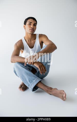 Barfuß-afroamerikaner in blauen Jeans, sitzt mit gekreuzten Beinen und sieht auf grauen Hintergrund in die Kamera, Stockbild Stockfoto