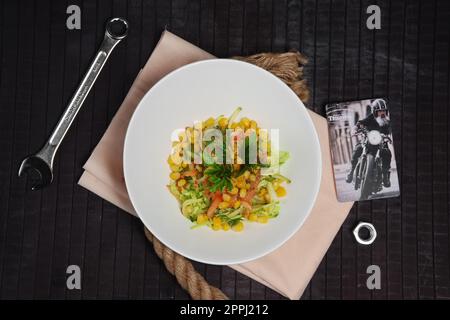 Salat mit Mais, Tomaten, Gurke und Kohl auf dem Tisch neben Schraubenschlüssel und Schraubenmutter. Draufsicht. Stockfoto
