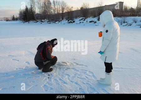 Logmozero, Karelien, Russland, 08. Januar 2022. Fischer und Fischer auf Winterfischerei. Ein Mann sitzt neben einem Loch in einem gefrorenen See und wartet auf einen Bissen. Schnee auf Eis, auf dem Boden nach Barsch fischen Stockfoto