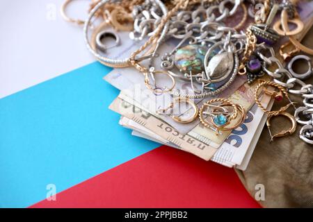 Ein Haufen gestohlener Schmuck und Geld auf Militäruniform Stoff und russische Flagge. Plünderung durch russische Soldaten in den ukrainischen Städten während des russischen Angriffs auf die Ukraine Stockfoto