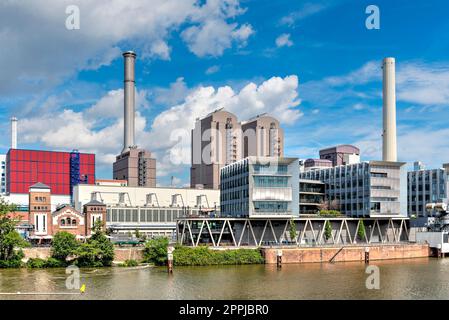 Das Ufer des Westhafens Frankfurt am Main mit Kraft-Wärme-Kopplungsanlage, Schornsteinen, Bürogebäuden und Industrieanlagen mit dem Main im Vordergrund bei Sommerwetter mit Frühlingswolken Stockfoto