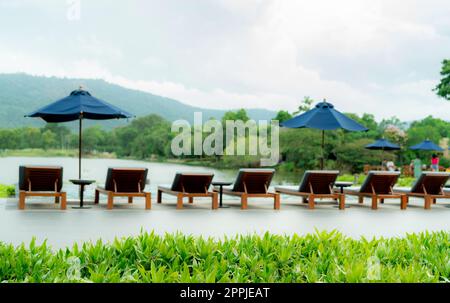 Grüner Rasen auf verwischtem Holzstrand und Sonnenschirm neben dem Swimmingpool in einem Luxushotel in der Nähe des Berges. Sommerurlaubsreisen. Tropischer Urlaub. Umweltfreundliches und nachhaltiges Hotel oder Resort Stockfoto