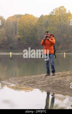 Porträt eines Reisenden am See im Herbst, der ein Foto mit der Digitalkamera und der orangefarbenen Jacke machte Stockfoto