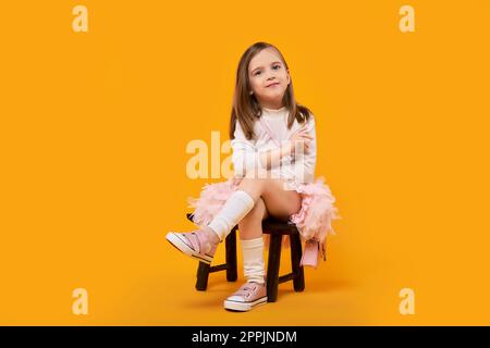 Junges Mädchen in Tüllrock und weißen Pullover sitzen auf Kleiner Holzhocker auf hellgelbem Hintergrund Stockfoto