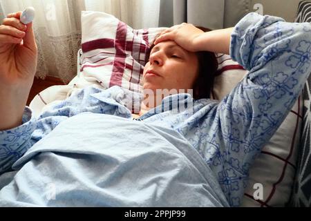 Kranke Frau im Bett. Kopfschmerzen, laufende Nase und Erkältung. Blässe des Gesichts und Ohnmacht des Körpers. Unwohlsein. Der Patient liegt auf einem Kissen, hält ein Thermometer und misst die Temperatur. Stockfoto