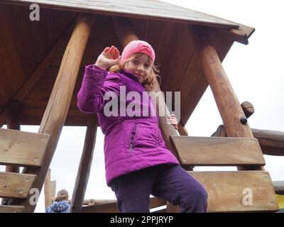 Mädchen 7 Jahre alt auf dem Spielplatz. Das Kind trägt warme Kleidung für die Halbzeit - eine lila Jacke und eine pinkfarbene Mütze. Holzspielplatz zum Klettern. Ein Mädchen winkt mit der rechten Hand Stockfoto