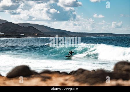 Wilde felsige Küste des Surfspots La Santa Lanzarote, Kanarische Inseln, Spanien. Surfer reiten eine große Welle in felsiger Bucht, Vulkan Berg im Hintergrund. Stockfoto