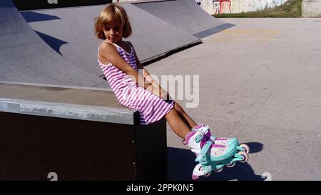 Sremska Mitrovica, Serbien, 12. September 2020. Das Mädchen fährt Rollerblading auf dem Asphalt. Ein 7-jähriges Kind in einem gestreiften weißen und pinkfarbenen Kleid fährt auf Rollschuhen auf dem Spielplatz. Stockfoto