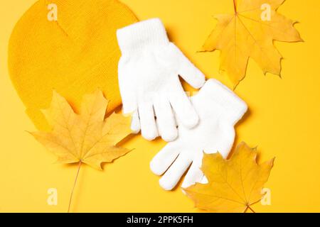 Stilvolle weiße Wollhandschuhe, Hut und trockene Blätter auf gelbem Hintergrund, flach liegend Stockfoto