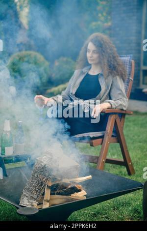 Brennholz im Grill im Freien und Unschärfe-Effekt auf junge Frau mit einem Glas Limonade im Hintergrund Stockfoto