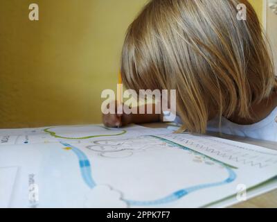 Ein 6-7-jähriges Mädchen liest ein Buch oder Lehrbuch und lächelt. Das Kind hat blonde Haare, dunkle Haut und Brille im Gesicht. Das Kind sitzt am Schreibtisch. Gelbe Wand im Hintergrund Stockfoto