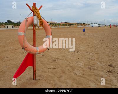 Anapa, Russland, 10. August 2021 Lifebuoy mit einer roten Flagge an einem Sandstrand. Orangefarbenes Rettungsboot an einer Stange, um Menschen zu retten, die im Meer ertrinken. Rettungspunkt am Ufer. Stürmisches Wetter am Schwarzen Meer Stockfoto